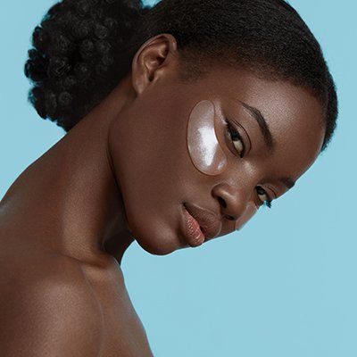 Close up portrait of beautiful dark-skinned african girl with reusable under eye makeup shield and silicone cooling pad used for reducing puffiness and helping with perfect makeup application. Young woman looking at camera. Concept of face skin care. Isolated on blue background. Studio shoot.