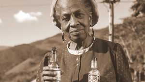 Ambrozine 'Adella' Kelly holding two bottles of her black castor seed oil shampoo in the scenic hills of Mile Gully, Saint Mary, Jamaica (circa 1948). Adella, a midwife and passionate beautician, stands proudly with her innovative hair care products, reflecting her dedication to promoting healthy hair growth and nourishment. The lush greenery of the Jamaican landscape serves as a backdrop, symbolizing the natural origins of Kintone's luxurious hair care line.