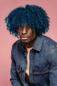 Stylish young black man with curly blue hair, rocking a denim jacket, poses confidently against a vibrant pink wall.